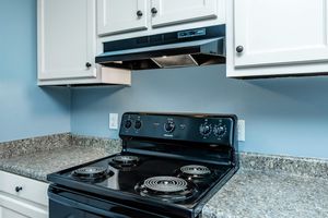 A black stovetop with four burners and knobs for temperature control, set against a light blue wall. Above it, there's a black ventilation hood installed beneath white cabinets, with a countertop made of speckled gray granite. The overall kitchen design is modern and clean.