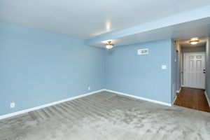 Empty room with light blue walls and beige carpet. A ceiling light fixture is visible, and there is a door on the right leading to an entryway. The space is well-lit and has a clean, minimalistic appearance.