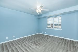 Empty room with light blue walls and a ceiling fan. The floor is covered with tan carpeting that has visible imprints. A window with white blinds allows natural light into the space. The room appears clean and freshly painted, suggesting it's ready for new occupants or furnishing.