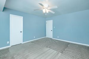 A minimalist bedroom with light blue walls and beige carpet. The room features a ceiling fan with five blades, two closed white doors on opposite walls, and no furniture, creating a spacious and empty atmosphere.