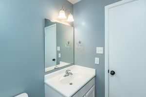 A small bathroom featuring a white sink with a chrome faucet and a large rectangular mirror above it. The walls are painted a light gray, and there are two light fixtures above the mirror. A white door and towel ring are visible, adding to the simple and clean design of the space.