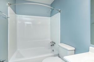 A clean, modern bathroom featuring a white bathtub with a curved shower rod, a white toilet, and blue-colored walls. The space is well-lit and tidy, showcasing a minimalist design with no visible clutter.