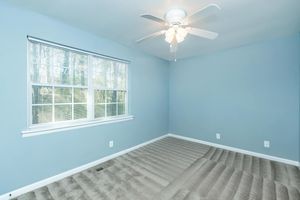 A spacious, light blue bedroom featuring a ceiling fan and a large window with natural light filtering in. The floor is covered with beige carpet, and the walls are simply decorated, creating a calm and serene atmosphere. The room appears freshly painted and clean.