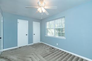 A clean, empty bedroom featuring light blue walls, a ceiling fan with light, and two white doors. A window allows natural light to enter, casting soft shadows on the beige carpeted floor. The room is well-kept and spacious, with a minimalist aesthetic.