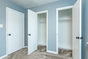 A light blue room featuring two open closet doors on the right and a closed door on the left. The closets are empty, showcasing white shelving inside. The floor is carpeted in a neutral tone, and the walls are painted a soft blue. The doors have simple black knobs.