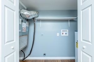 A clean and well-lit laundry room featuring an HVAC unit on the left, a white shelf mounted on the wall, and a small utility access panel. The walls are painted gray, and the flooring is wood. Two open white closet doors frame the space, providing easy access.