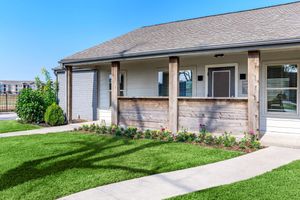 A well-maintained suburban home with a front porch featuring wooden posts. Neatly trimmed grass surrounds the pathway leading to the entrance. Colorful flowers line the porch, contributing to the inviting atmosphere. The sky is clear and blue, enhancing the bright appearance of the property.