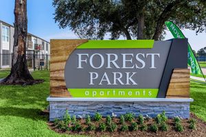 Signage for Forest Park Apartments, featuring a modern design with the words "FOREST PARK" prominently displayed in large letters. The background includes a combination of wood and stone textures, with green accents. Surrounding the sign are well-maintained lawns and flower beds.