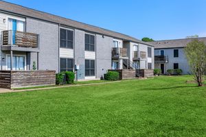 Two multi-unit residential buildings with gray exteriors and wooden balconies situated in a well-maintained green lawn. The buildings feature multiple windows, and there are small bushes and trees around the grounds under a clear blue sky.