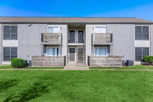 A two-story apartment building with a gray exterior. The building features two sets of stairs leading to upper-level balconies. Each unit has a front door with windows on either side. The lush green lawn in front is well-maintained, creating a welcoming outdoor space. Clear blue sky above enhances the bright appearance.