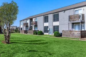 Two-story apartment buildings with gray brick exteriors, featuring balconies and surrounded by neatly trimmed greenery. A small tree is visible in the foreground, and the lawn is well-maintained, creating a peaceful residential environment under a clear blue sky.