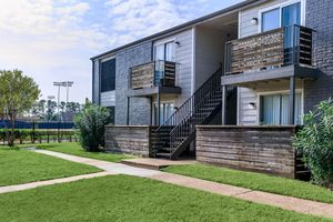 Two-story apartment building featuring a grey exterior with wooden balcony railings. Each unit has a staircase leading to the upper floor. Lush green grass surrounds the building, and there is a sidewalk leading up to the entrance. In the background, sports fields can be seen under a bright blue sky.