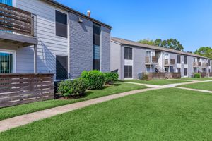 Two-story apartment buildings with gray and white exteriors surrounded by manicured, green lawns. Sidewalks lead through the grassy area, and balconies are visible on some units. Bright blue sky overhead creates a pleasant and inviting atmosphere.