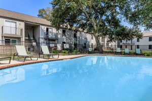 A clear blue swimming pool with lounge chairs around it, surrounded by landscaped areas and a few trees. In the background, there are multi-story apartment buildings with balconies and railings, set against a bright blue sky.