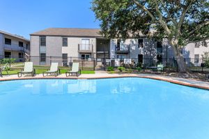 A clear blue swimming pool surrounded by green grass and lounge chairs. In the background, a multi-story apartment building with balconies is visible, under a bright blue sky and a large tree providing shade near the pool area.