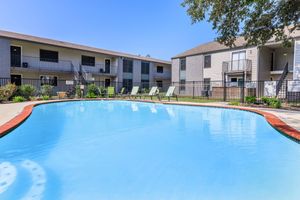 A clear blue swimming pool surrounded by lounge chairs, set among landscaped greenery. Two apartment buildings with balconies can be seen in the background, under a bright blue sky. The scene conveys a relaxed, inviting atmosphere ideal for leisure and outdoor enjoyment.
