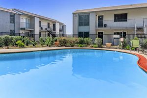 A clear blue swimming pool framed by green landscaping, with two multi-story residential buildings in the background. The pool area features a wooden deck and potted plants, creating a serene outdoor environment.
