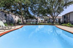A sparkling blue swimming pool surrounded by green trees and lounge chairs, with apartment buildings in the background. The scene conveys a sunny and inviting outdoor space, ideal for relaxation and leisure.
