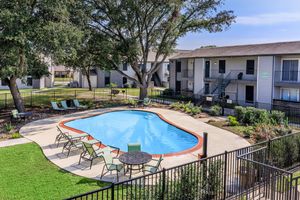 A serene outdoor swimming pool surrounded by chairs and a table, with lush greenery and trees nearby. Apartment buildings are visible in the background, creating a welcoming atmosphere. The scene is bright and inviting, perfect for relaxation and leisure.