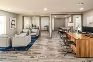 A modern office space featuring a reception area with white chairs and a blue rug. In the background, glass partitioned meeting rooms are visible. There is a wooden desk with black chairs, and large windows provide natural light. The decor is minimalistic and professional, creating a welcoming atmosphere.