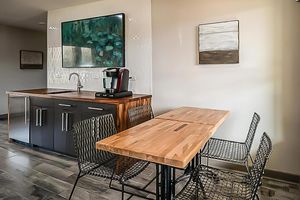 A modern kitchen space featuring a wooden table with black wire chairs. In the background, there's a kitchenette with a coffee maker, a sink, and art on the walls, including a large painting and a smaller piece. The decor combines wood and sleek dark finishes for a contemporary look.