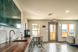 A modern interior of a lounge area featuring a wooden countertop with a sink, a coffee maker, and an empty seating area with two white chairs. Large windows let in natural light, and there is a flat-screen TV mounted on the wall. The space has a sleek, contemporary design with neutral colors.