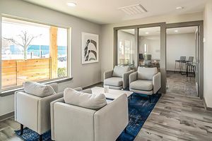 A modern, well-lit interior space featuring two gray armchairs and a coffee table on a blue area rug. A large window offers a view of the outdoors, while a glass-walled room in the background contains additional seating. The walls are painted a light color, creating a clean and inviting atmosphere.