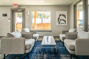 A modern lounge area featuring two light gray armchairs and a matching sofa around a white coffee table on a blue patterned rug. A large window provides natural light, framing a view of trees. The walls are neutral, with minimal artwork and an exit sign visible.