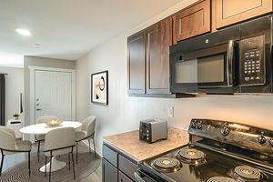 A modern kitchen featuring dark wood cabinets, a black microwave, and a sleek black stove. A small silver toaster is on the countertop. In the background, there is a dining area with a round white table and grey chairs, illuminated by natural light coming from a nearby door.