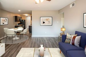 A modern living room featuring a navy blue sofa with decorative pillows, a coffee table with a vase, and a light-colored rug. In the background, there's a dining area with a round table and four chairs, surrounded by artwork on the walls. The kitchen is visible, showcasing dark cabinetry and appliances.