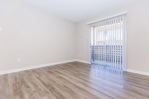 Empty room with light-colored walls and wooden flooring. A sliding glass door with vertical blinds leads to a small outdoor area, letting in natural light. The room is unfurnished, creating a spacious and open feel.