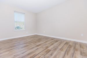 Empty room with light-colored walls and a large window with vertical blinds. The floor is made of wooden laminate, featuring a warm brown tone. The space is well-lit and devoid of furniture, creating a minimalist atmosphere.