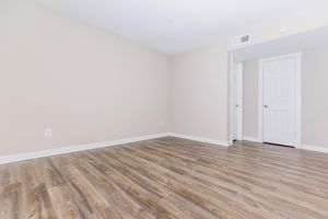 A spacious, vacant room with light beige walls and hardwood flooring. A single door is visible on the right, leading to another space, while the wall on the left features an electrical outlet. Natural light fills the area, creating an inviting atmosphere.