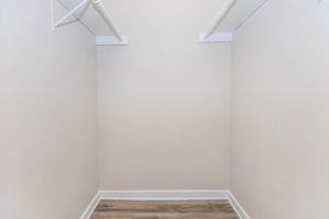 A vacant closet featuring two white clothing rods against plain beige walls. The floor is finished with light wood-like panels, and there is ample space for storage. The overall appearance is clean and minimalistic.