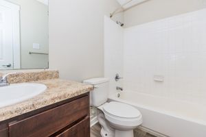 A bathroom featuring a bathtub with a showerhead, a toilet, and a granite countertop sink. The walls are painted a light color, and there is a large mirror above the sink. The flooring appears to be laminate.