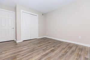 An empty room with light beige walls and wooden laminate flooring. There are white double doors to the left and a single white door on the far side, leading into a closet or another space. The room has a simple, clean aesthetic with no furniture or decorations.