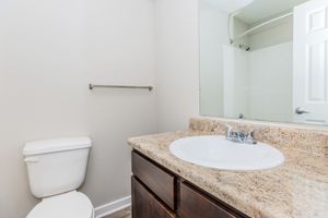 A clean bathroom featuring a white toilet, a granite countertop with a round sink, a wall-mounted mirror, and a towel rack. The space includes light-colored walls and a shower area visible in the background. The overall design is modern and minimalistic.