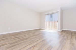 A spacious, unfurnished room with light-colored walls and a large window featuring vertical blinds. The room has a wooden floor and natural light streaming in from the window, providing a bright and airy atmosphere.