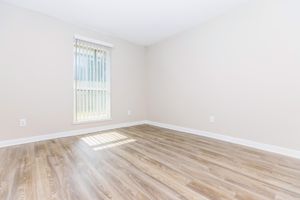 A bright, empty room with light-colored walls and faux wood flooring. A single window with vertical blinds allows natural light to enter, casting shadows on the floor. The space is clean and minimalist, suitable for various uses or decoration. No furniture or other items are present.
