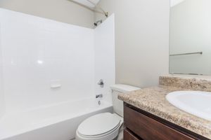 A clean bathroom featuring a white bathtub with a shower, a toilet, and a granite countertop with a sink. The walls are painted in a soft beige color, and there is a mirror above the sink. The overall design is modern and minimalistic, providing a bright and spacious feel.