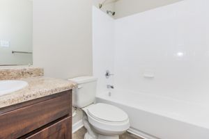 A clean and simple bathroom featuring a bathtub with a shower, a toilet, and a dark wood vanity with a granite countertop. The walls are light-colored, and there's a mirror above the sink reflecting the space. The overall design is modern and minimalistic.