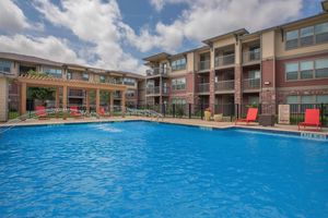 A vibrant blue swimming pool surrounded by lounge chairs and a shaded pergola. In the background, there are modern apartment buildings with balconies and large windows. The scene is bright with a partly cloudy sky.