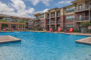 A bright blue swimming pool surrounded by lounge chairs and a shaded pergola. Modern apartment buildings in the background feature balconies and greenery. The sky is partly cloudy, creating a relaxing outdoor atmosphere.
