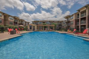 A clear blue swimming pool surrounded by lounge chairs in a residential complex. In the background, there are modern apartment buildings with landscaped greenery and a partly cloudy sky. The scene conveys a relaxing outdoor space for residents.