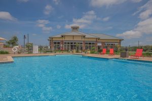 A clear blue swimming pool surrounded by lounge chairs, with a two-story building in the background. The setting features a well-maintained landscape, with a serene sky and a few clouds. The pool area is enclosed by a fence, enhancing the inviting atmosphere.