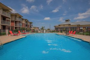 A clear blue swimming pool in an outdoor setting, surrounded by lounge chairs in bright red. Nearby, there are buildings with balconies. The sky is partly cloudy, and there's a clubhouse visible in the background, creating a vibrant and inviting atmosphere.