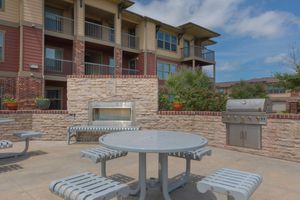 Outdoor patio area featuring a circular table with attached benches, a built-in stone grill, and an outdoor fireplace. Surrounding greenery adds a touch of nature, while two multi-story apartment buildings with balconies are visible in the background. The sky is partly cloudy.