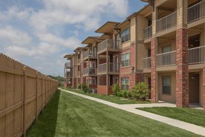 A row of modern, multi-story apartment buildings with balconies, set alongside a manicured green lawn. A wooden fence runs parallel to the walkway, which leads past the buildings. The sky above is partly cloudy, creating a bright and inviting atmosphere.