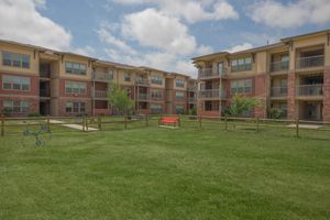 A well-maintained residential complex with multiple buildings featuring balconies. The grassy courtyard has a red bench and a fenced area, along with some small trees and a decorative structure resembling a spinning lawn ornament. The sky is partly cloudy.