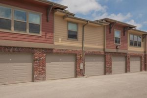 A row of residential buildings featuring a mix of brick and siding exteriors. The lower levels have garage doors, while the upper levels have windows. The sky is partly cloudy, and the ground is paved, indicating a well-maintained neighborhood or apartment community.